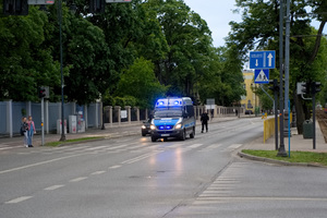 policyjny radiowóz z włączonymi sygnałami świetlnymi stoi przy skrzyżowaniu. Obok radiowozu przechodzi policjant