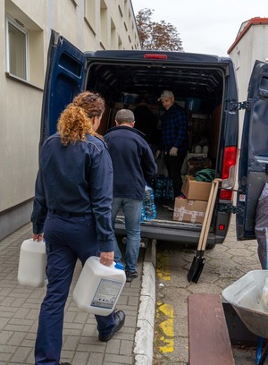 Policjantka idzie w kierunku samochodu dostawczego niosąc dwa baniaki z białym płynem