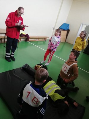 Sala gimnastyczna. Zawodnicy udzielają pomocy poszkodowanemu. Ocenia to ratownik medyczny.