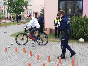 Teren przy szkole. Zawodnik pokonuje slalom na rowerze. Ocenia to policjant