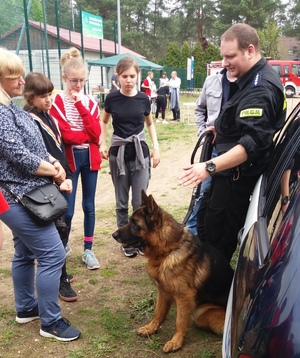 Zewnętrzny teren szkoły. Policjantka- przewodnik psa służbowego rozmawia z uczestnikami konkursu. Towarzyszy temu pies policyjny.