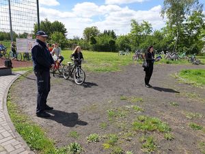 Teren zielony przy szkole. Policjant nadzoruje egzamin na kartę rowerową. W tle inni zdający.