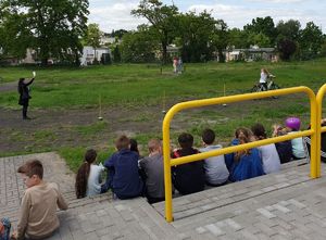 Teren zielony przy szkole. Uczennica podczas egzaminu pokonuje tor rowerem. Przyglądają się temu inni uczniowie.