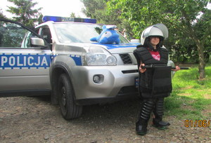 Dziecko stojąc przed radiowozem pozuje do zdjęcia przebrane w strój policjanta.