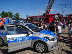 Policyjny radiowóz stojący na tle pojazdu straży pożarnej. Stojąca przy nim policjantka pokazuje dzieciom i dorosłym &quot;magiczną&quot; tubę.