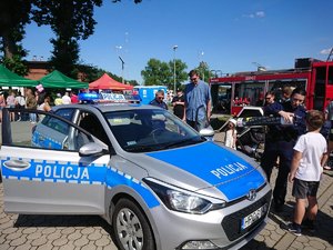 Policyjny radiowóz stojący na tle pojazdu straży pożarnej. Stojąca przy nim policjantka pokazuje dzieciom i dorosłym &quot;magiczną&quot; tubę (2).