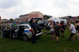 Nastolatkowie oglądają policyjną tarczę, hełm oraz pałkę typu tonfa. Przygląda się temu policjant. W kadrze znajduje się również policyjny radiowóz.