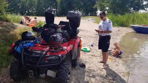 Policjant podczas kontroli Quada zaparkowanego na plaży tuż przodzie. Obok bawiące się dziecko, a w tle osoby odpoczywające na wodą.