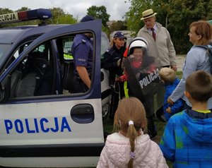 Policjanci przy radiowozie rozmawiają z uczestnikami festynu. Wśród nich stoi chłopiec, który ma założone elementy policyjnego wyposażenia, m. in. kamizelkę kuloodporną, hełm i tarczę.