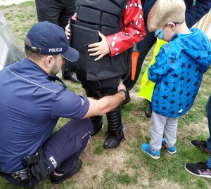 Policjant zakłada chłopcu elementy policyjnego uzbrojenia szturmowego- kamizelkę kuloodporną oraz ochraniacze na nogi. Przyglądają się temu inne dzieci.