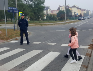 Dwie uczennice pod okiem policjanta przechodzą przez przejście dla pieszych.