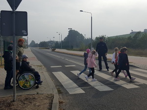 Dzieci wraz z opiekunem pod okiem policjanta pokonują przejście dla pieszych.