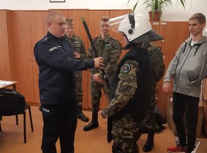 Policjant zakłada jednemu z uczniów strój policyjny bojowy. Przyglądają się temu pozostali uczniowie.