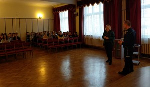 Policyjni profilaktycy podczas zajęć. W tle siedzący uczniowie.