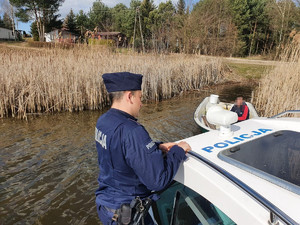 Policjant stojąc na pokładzie policyjnej łodzi motorowej rozmawia z wędkarzem na łódce.