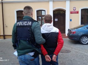 Policjant prowadzi jednego z zatrzymanych sprawców. W tle budynek prokuratury.