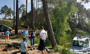 Policjant rozmawia z mężczyznami, którzy wjechali pojazdami do lasu. W tle również klika innych osób w różnym wieku.