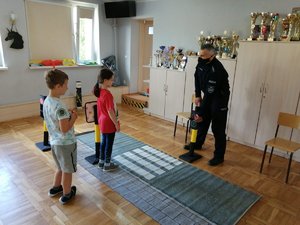 Zajęcia praktyczne w sali placówki. Policjant omawia z dziećmi bezpieczne przechodzenie przez jezdnię przy użyciu miasteczka ruchu drogowego.