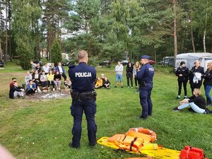 Policjanci rozmawiają z uczniami  siedzącymi na ławkach i trawie.