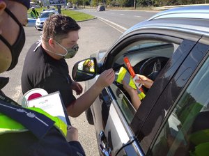 Pracownik PZMotu wręcza kierowcy materiały profilaktyczne. Obok stoi policjant.