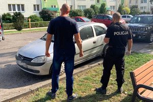 dwaj policjanci stoją przed skradzionym srebrnym oplem corsa zaparkowanym na ulicy