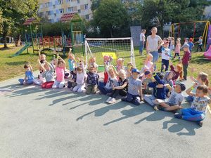 Dzieci siedząc w półkolu unoszą w górę rękę z założoną opaską odblaskową.