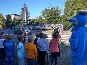 Dzieci wraz z policjantami i polfinkiem stoją przy przejściu dla pieszych przy pobliskiej ulicy.