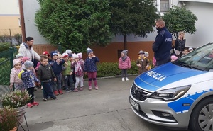 Dzieci  z wychowawczynią stoją przed zaparkowanym radiowozem. Obok stoi policjant.