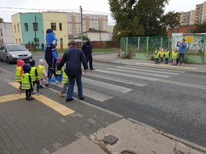 Dzieci stoją po obu stronach przejścia dla pieszych i czekają na przejście pod okiem policjantów i wychowawców.