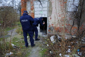 Policjanci przy pustostanie. jden z lich świecąc latarką patrzy do środka przez otwór w ścianie.