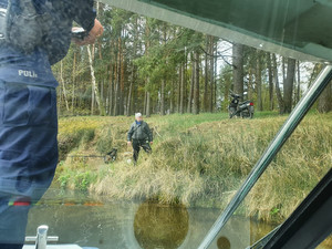 Policjant na łodzi motorowej. W tle las i woda. Na brzegu stoi mężczyzna łowiący ryby. Obok niego widoczny zaparkowany motocykl w lesie.
