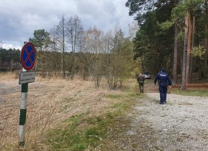 Policjant i leśnik idą w stronę zaparkowanego przy brzegu samochodu osobowego widocznego w głębi zdjęcia. po lewej stronie widoczny zakaz zatrzymywania i postoju.