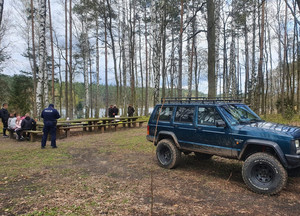 Policjant i leśnicy rozmawiają z osobami siedzącymi na parkingu leśnym na ławkach. Po prawej stronie zdjęcia widoczny zaparkowany samochód terenowy.
