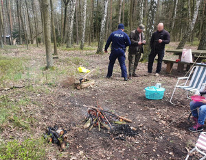 Policjant rozmawia z dwoma mężczyznami. Przed nimi widoczne dwa rozpalone ogniska.