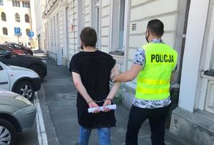 Policjant prowadzi zatrzymanego po chodniku. Z lewej strony widoczne zaparkowane pojazdy, a z prawej budynek.