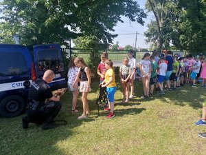 Dzieci stoją w kolejce do pogłaskania psa policyjnego. Przed nimi kuca policjant trzymający psa policyjnego.