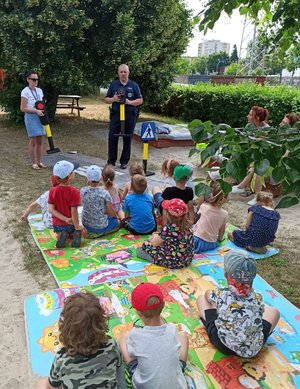 Policjant prowadzi zajęcia z dziećmi na terenie zielonym przedszkola. Dzieci siedzą na macie i słuchają prelekcji policjanta.