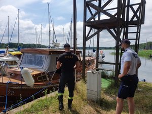 Policjant i strażak stoją przed łodzią przycumowaną do nabrzeża.