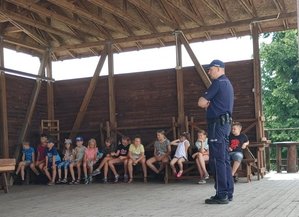 Uczestnicy podczas spotkania. Dzieci siedzą na ławce pod zadaszeniem. Przed nimi stoi policjant.