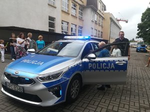 Policjant stoi przy radiowozie. Za nim stoją wychowawczynie z dziećmi.