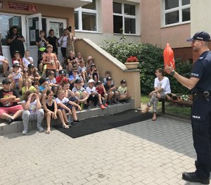 Policjant prezentuje dzieciom bojkę ratowniczą. Dzieci siedzą na schodach szkoły.