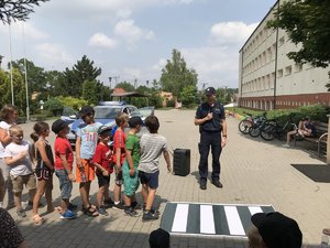 Policjant i kilkoro dzieci stoją przed matą imitującą  przejście dla pieszych leżącą na placu .