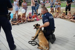 Policjant z psem służbowym rozmawia z dziećmi siedzącymi przed nim w półkolu.