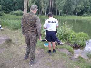 Policjant i leśnik podczas legitymowania wędkarza siedzącego nad brzegiem wody i łowiącego ryby.