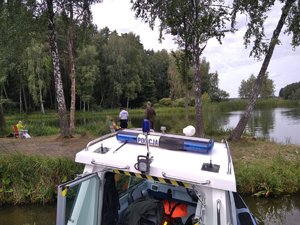 Na pierwszym planie część policyjnej łodzi motorowej, a w tle policjant i leśnik rozmawiają z wędkarzami siedzącymi nad brzegiem akwenu.