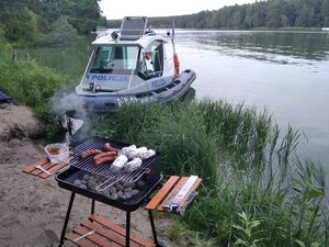 Palący się grill z kiełbaskami. W tle policyjne łódź motorowa na wodzie.