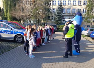 Policjanci wraz z Polfinkiem rozmawiają z dziećmi przed szkołą.