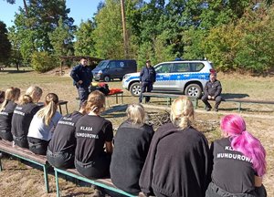 policjanci i uczniowie podczas zajęć prowadzonych w terenie zielonym. Uczestnicy siedzą na ławkach ustawionych w kwadrat.