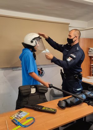 Policjant zakłada Maksymilianowi elementy policyjnego umundurowania ćwiczebnego, w tym kask.