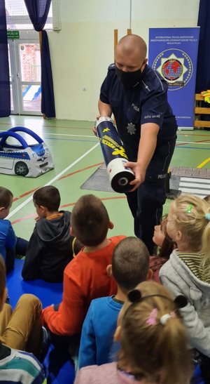 Policjant prezentuje dzieciom magiczną tubę, przez którą można porównać jak widoczna jest osoba która ma na sobie odblaski, z tą która nie ma.